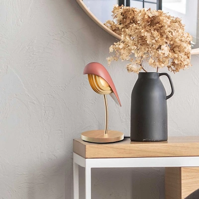 Auf einem Sideboard mit weissen Beinen und Holzplatte steht die Bird-Lampe aus rosa Porzellan mit goldenen Akzenten und Fichtenholz Sockel. Daneben eine schwarze Vase mit beigen Trockenblumen. Dahinter eine grau strukturierte Wand mit rundem Spiegel