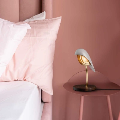 Ein sanftes, rosa Ambiente umh&uuml;llt ein Bett mit weisser Bettw&auml;sche und zartem, hellrosa Kissen. Ein rosa Tischchen tr&auml;gt die warm leuchtende Bird-Lampe in Almond Beige mit goldenen Akzenten und schafft eine ruhige, behagliche Stimmung.