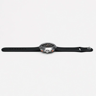 Seitliche Ansicht einer Armbanduhr mit schwarzem Silikonarmband auf wei&szlig;em Hintergrund. Das runde Metallgeh&auml;use zeigt ein dunkles Zifferblatt, seitlich sind Krone und zwei orangefarbene Dr&uuml;cker sichtbar. Minimalistische Produktaufnahme. Sehr sachlich.